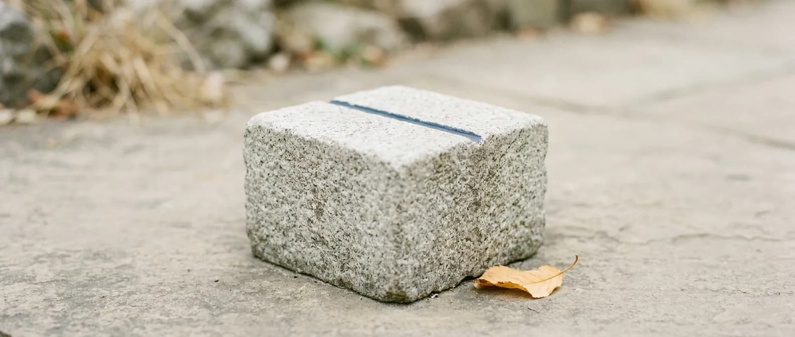 Ein kleiner verwitterter Grenzstein aus hellgrauem Granit steht auf einer hellen Fläche, die Oberseite leer bis auf eine flache dunkelblaue Rille, daneben ein trockenes gelbes Blatt