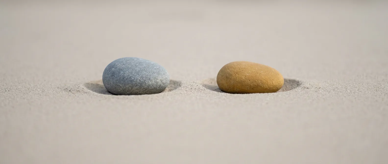 Zwei glatte Flusskiesel liegen mit Abstand nebeneinander auf hellem trockenem Sand, einer kühl grau, einer warm ockergelb