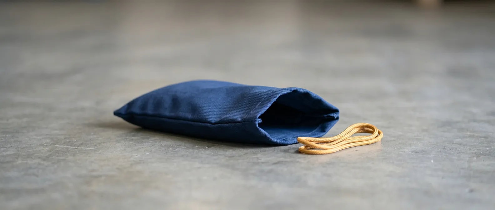 Ein leerer, nie gefüllter dunkelblauer Sandsack liegt flach zusammengelegt auf einem hellen Betonboden, daneben eine kurze gelbe Kordel