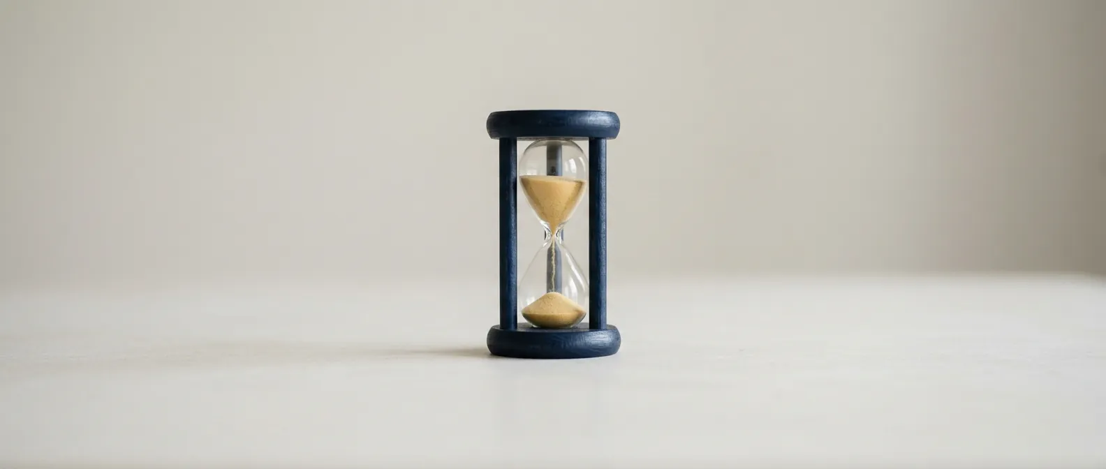 Eine kleine Sanduhr mit dunkelblauem Holzrahmen steht allein auf einer hellen Fläche, etwa die Hälfte des hellgelben Sandes ist durchgelaufen, der Rest rieselt noch