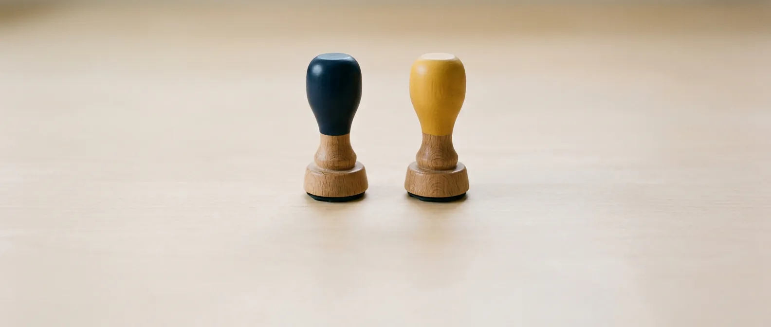 Zwei gleiche Holzstempel stehen aufrecht nebeneinander auf einem hellen Tisch, ein Griff dunkelblau, einer gelb, die Stempelflächen nach unten gedreht
